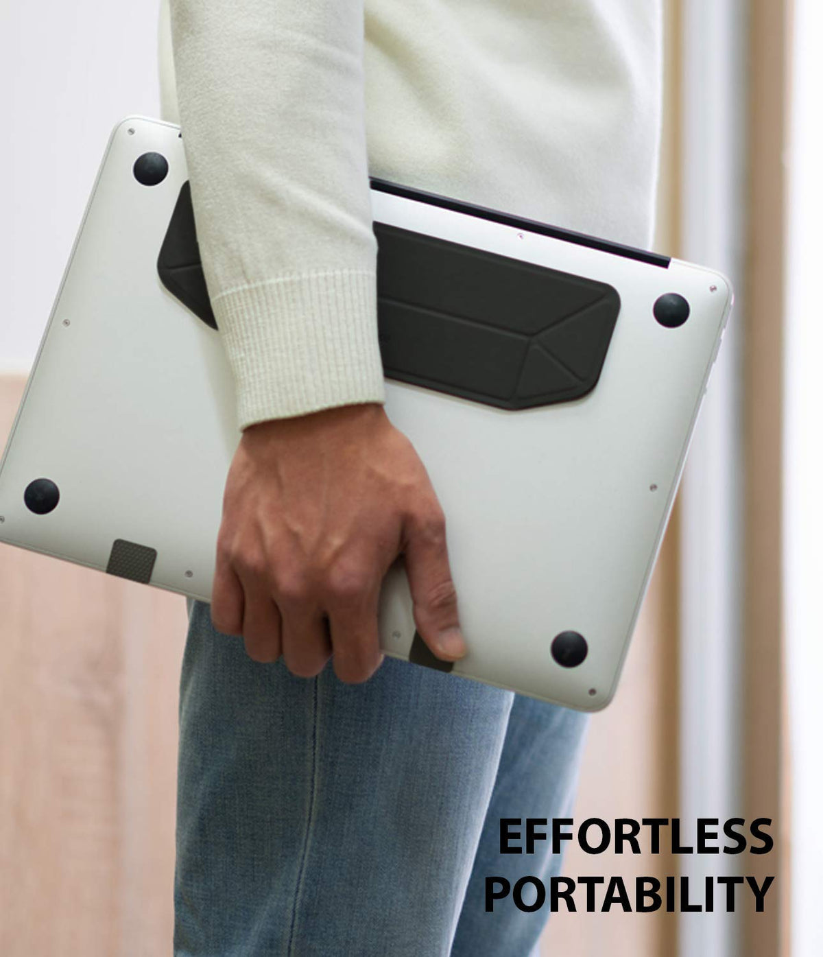 Laptop Stand - Person carrying a silver laptop with a slim, black adhesive stand attached to the bottom.