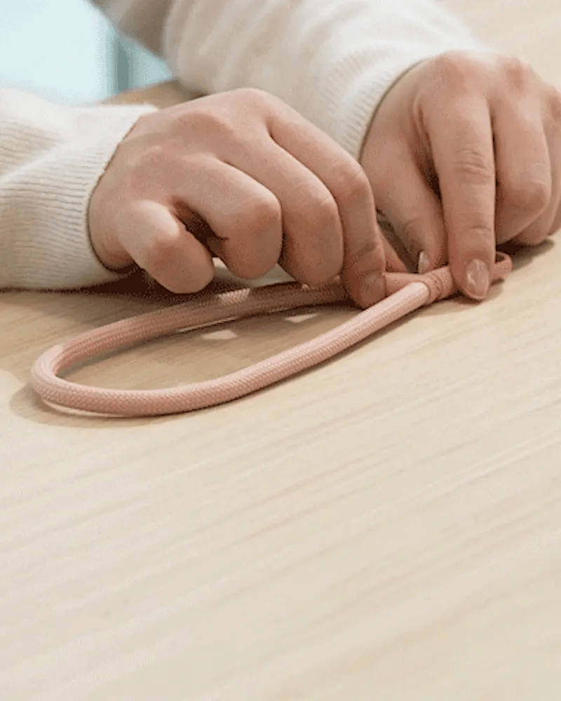 Holder Link Flow Hand Strap P-Type - Hands adjusting a looped pink woven cord strap on a light wooden surface.