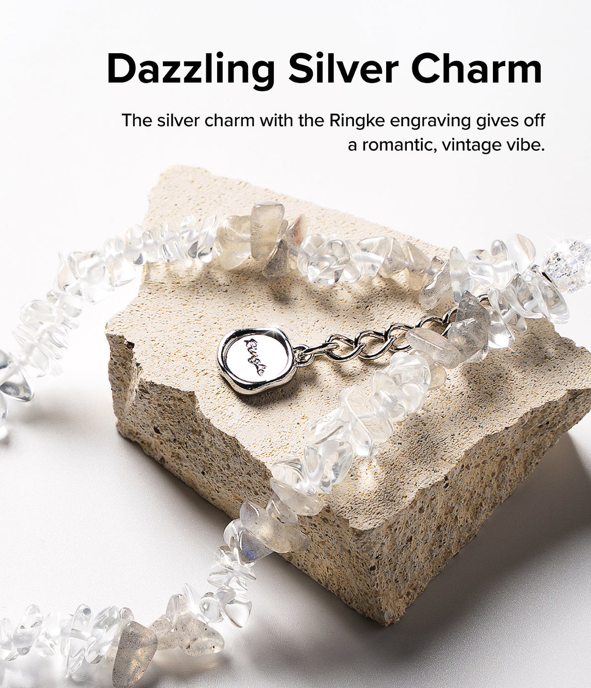 Beaded Strap - A silver charm with Ringke engraving on a clear crystal beaded strap resting on a textured stone block.