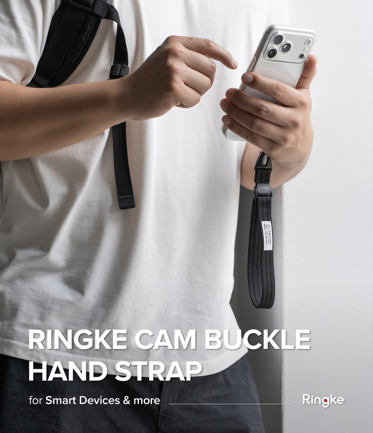 Cam Buckle Hand Strap - A person wearing a white t-shirt holds a silver smartphone with a black Ringke camera buckle hand strap attached. The person is pointing at the strap.