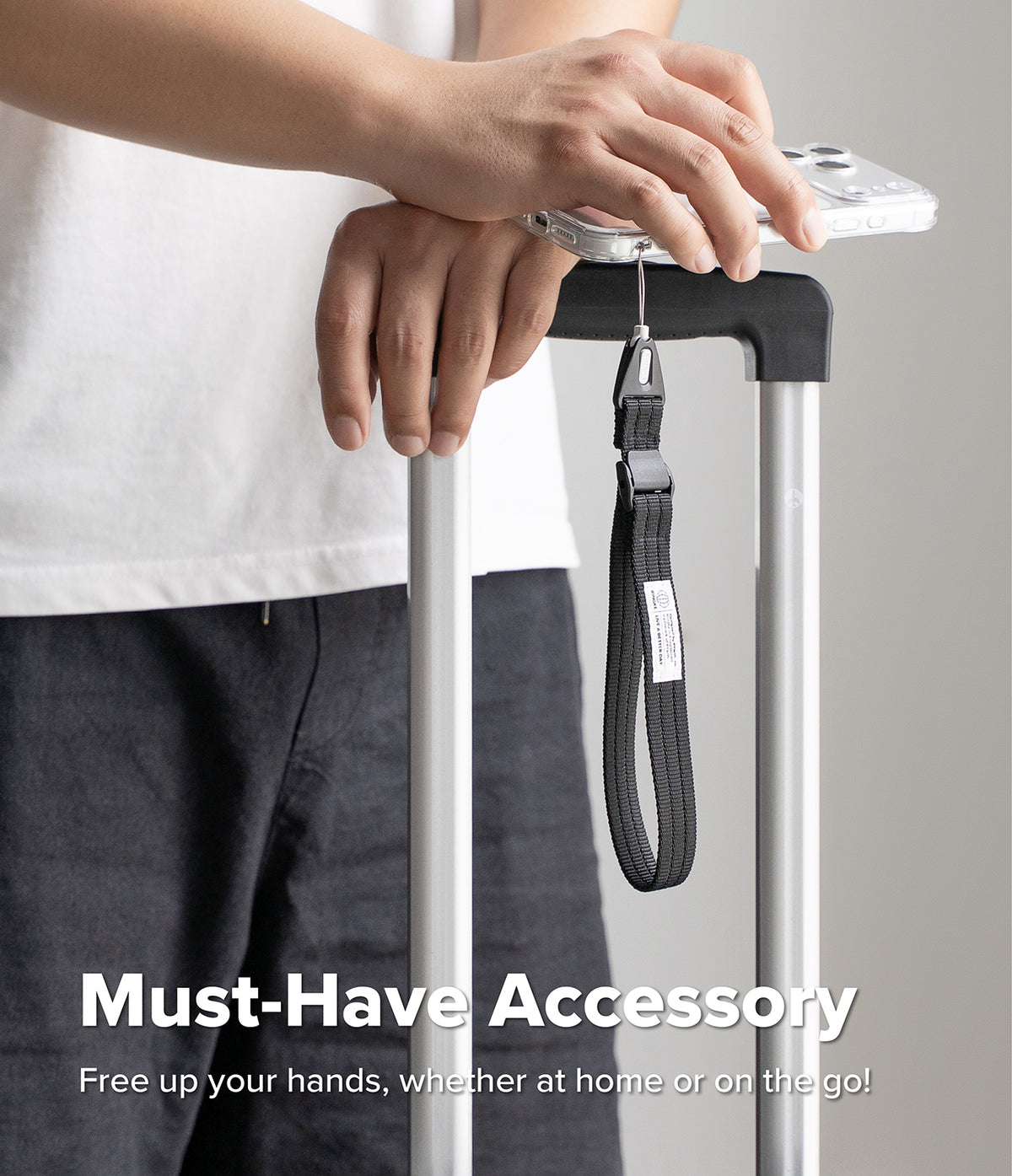 Cam Buckle Hand Strap - A close-up of a person holding a silver rolling suitcase handle with a black wrist strap attached to the top, with a phone resting on the handle. The person is wearing a white t-shirt and dark gray pants.