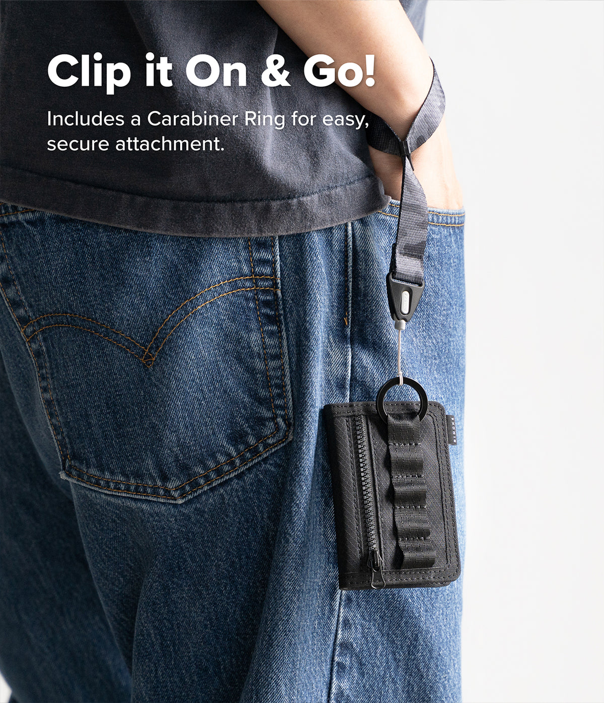 Wallet | Tech Magnetic - A close-up of a person wearing blue jeans and a dark gray shirt, showing a black zippered wallet or card holder clipped to the back pocket with a strap and ring. White text at the top reads 'Clip it On & Go! Includes a Carabiner Ring for easy, secure attachment.