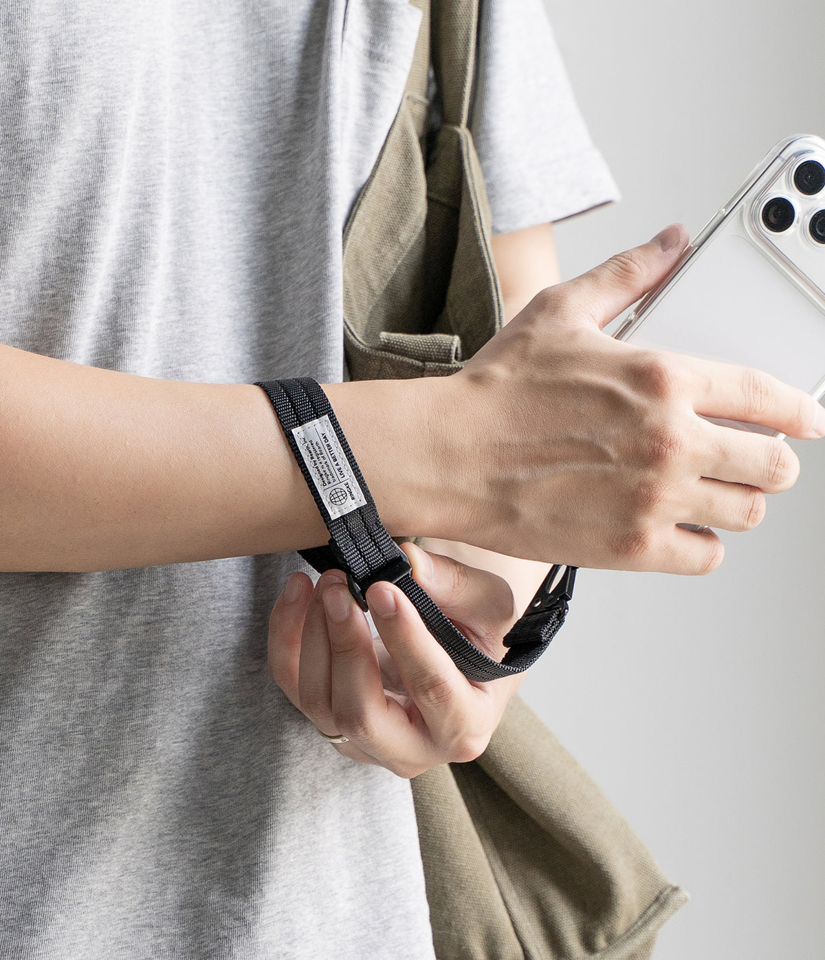 Holder Link Tactical Cam Buckle Hand Strap - A close-up of a person wearing a black adjustable wrist strap with a white label, holding a white smartphone and a khaki shoulder bag.