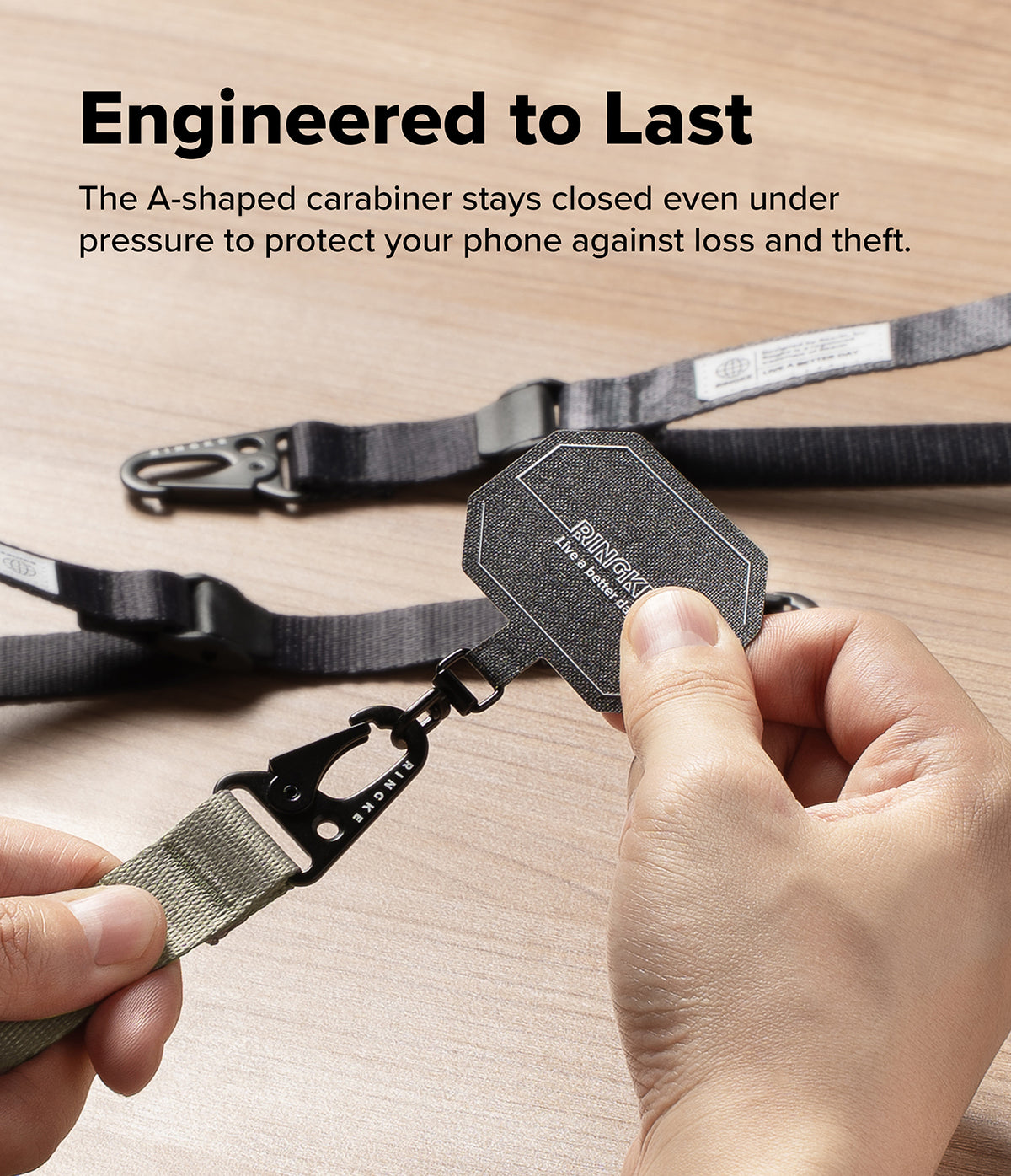 Holder Link Tactical Cam Buckle Hand Strap - A close-up of a person holding a black octagonal carabiner with a gray webbed strap, with more of the same straps laid out on a light wood surface. Text at the top reads, 'Engineered to Last' and describes the carabiner staying closed under pressure.