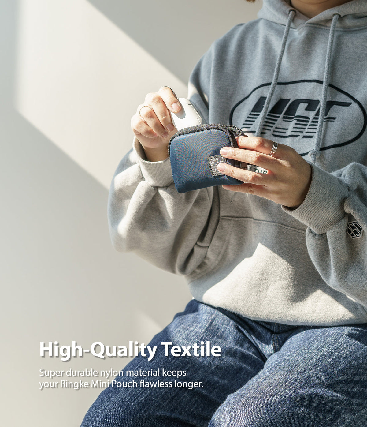 Mini Pouch | Half-Pocket - Person in a grey hoodie placing white wireless earbuds into a small blue nylon zippered pouch.