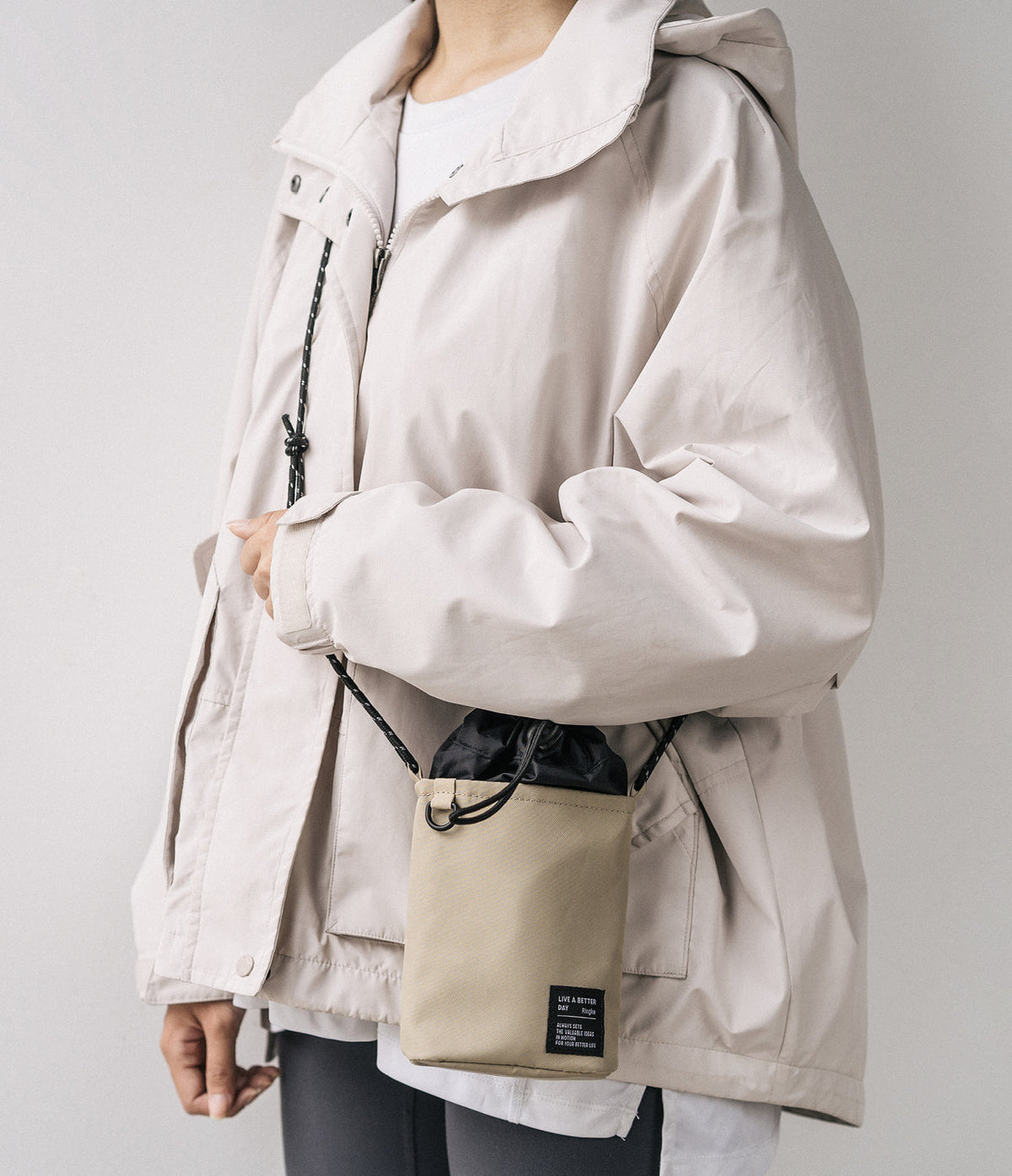 Mini Cross Bag | Bucket Bag - Person wearing a beige jacket and a small beige bucket bag with a black drawstring closure.