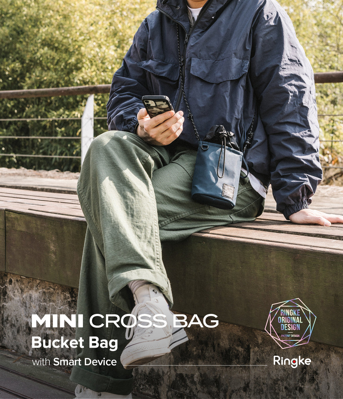 Mini Cross Bag | Bucket Bag - Person wearing a navy blue bucket bag crossbody while sitting on a bench holding a smartphone.