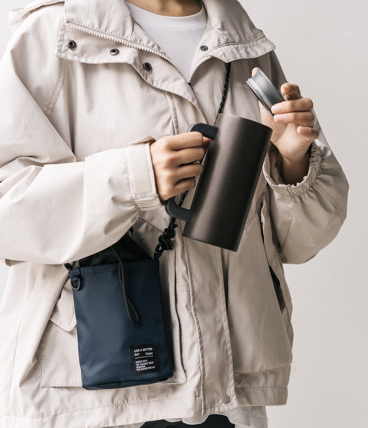 Mini Cross Bag | Bucket Bag - Person wearing a beige jacket and a navy blue bucket bag, holding a dark travel mug.