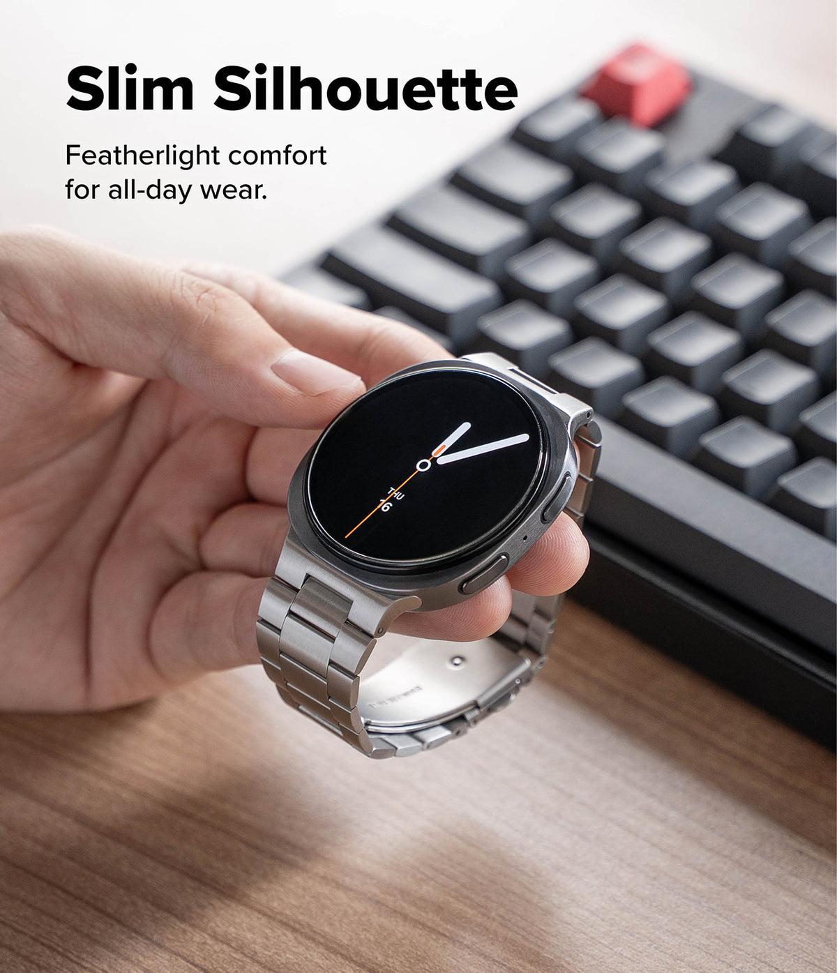 Galaxy Watch 9 / 8 | Metal One Air - A hand holds a slim round smartwatch with a silver metal link band and a black display, with a blurred keyboard and desk surface in the background. Text at the top reads 'Slim Silhouette' and 'Featherlight comfort for all-day wear.