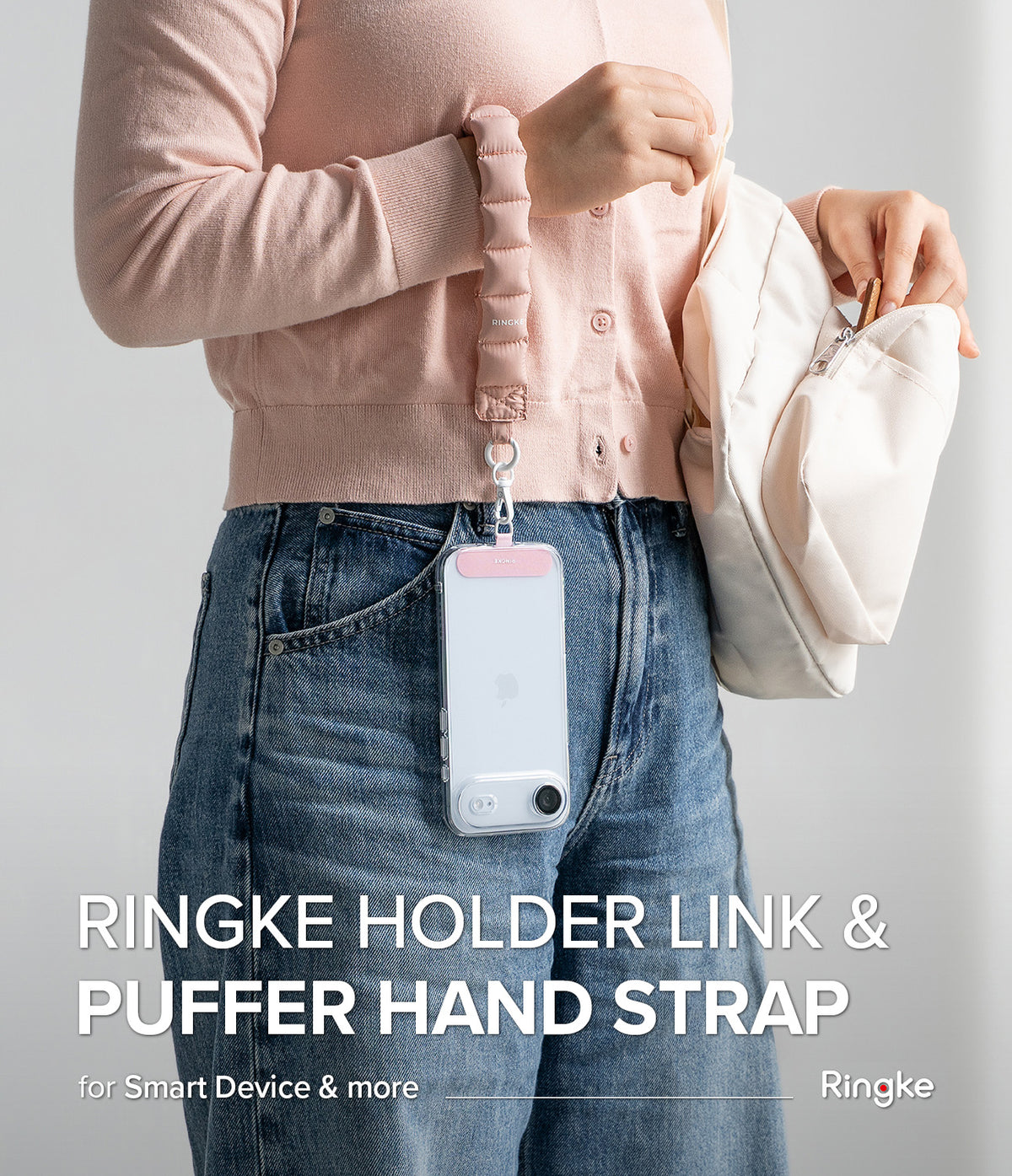 Holder Link Puffer | Hand - A person wearing a pink puffer wrist strap attached to a clear phone case hanging against blue jeans.