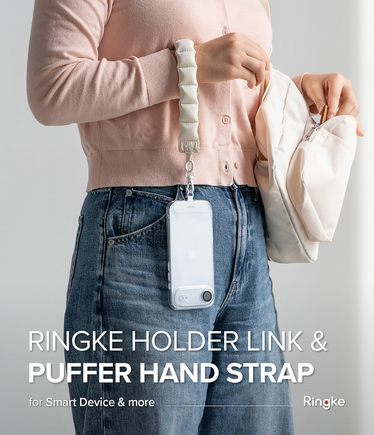 Holder Link Puffer | Hand - A person wearing a beige puffer wrist strap attached to a clear phone case hanging against blue jeans.
