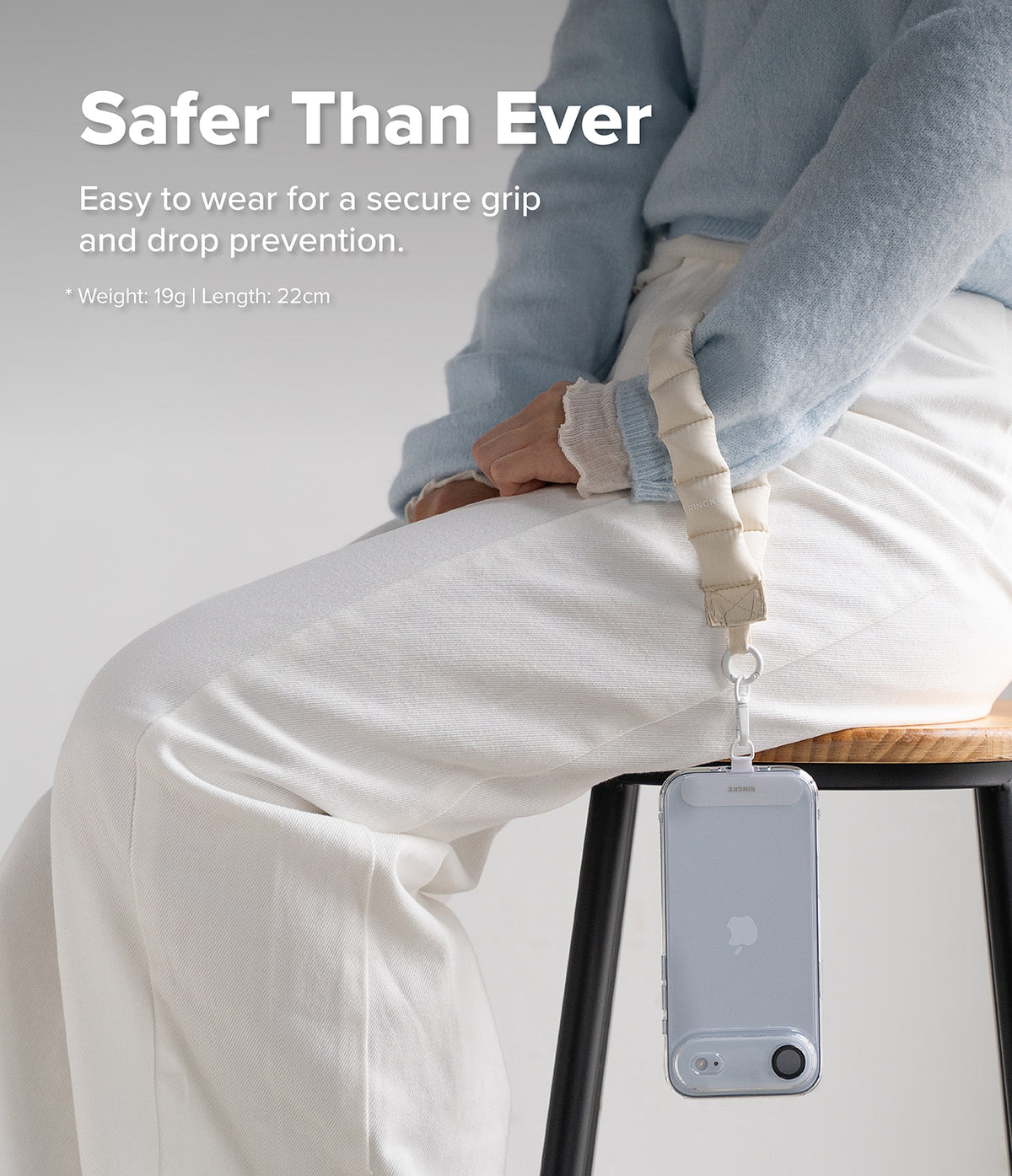 Holder Link Puffer | Hand - Person wearing a beige quilted puffer wrist strap attached to a clear phone case while sitting.