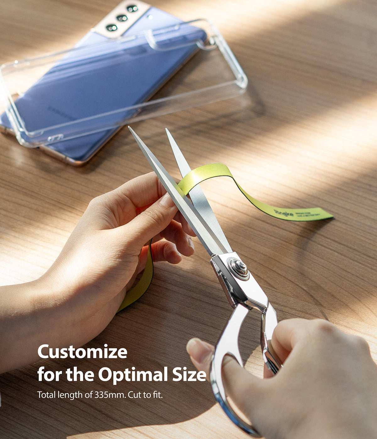 Ringke Strap Band - Hands cutting a yellow strap with scissors on a wooden table next to a smartphone in a clear case.