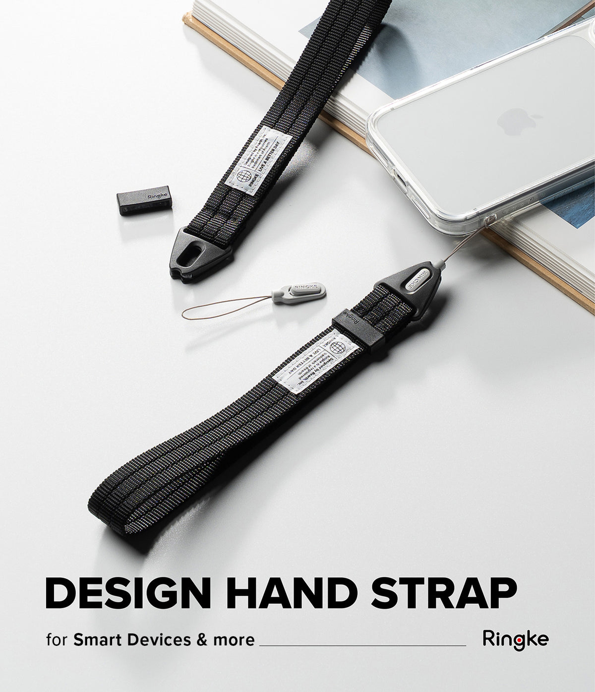 Hand Strap | Basic - A black woven hand strap with a metal clip and a small attached lanyard tag, shown alongside a smartphone and books on a light gray surface. Large text at the bottom reads 'DESIGN HAND STRAP' with smaller text 'for Smart Device & Every things' and the brand name 'Ringke' at the lower right.