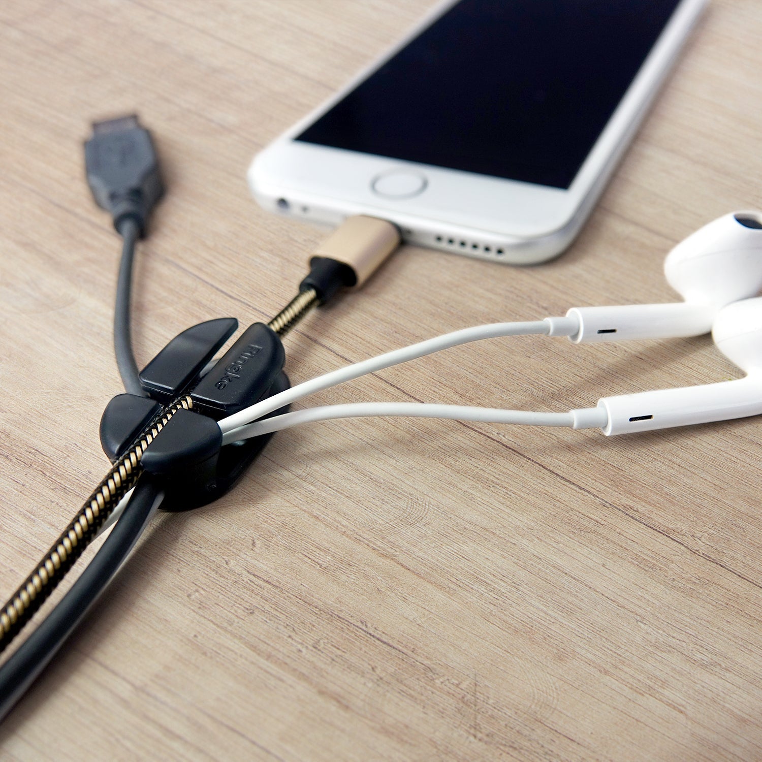 Cable Holder - A black cable organizer securing a charging cable and white earphones on a wooden desk next to a smartphone.