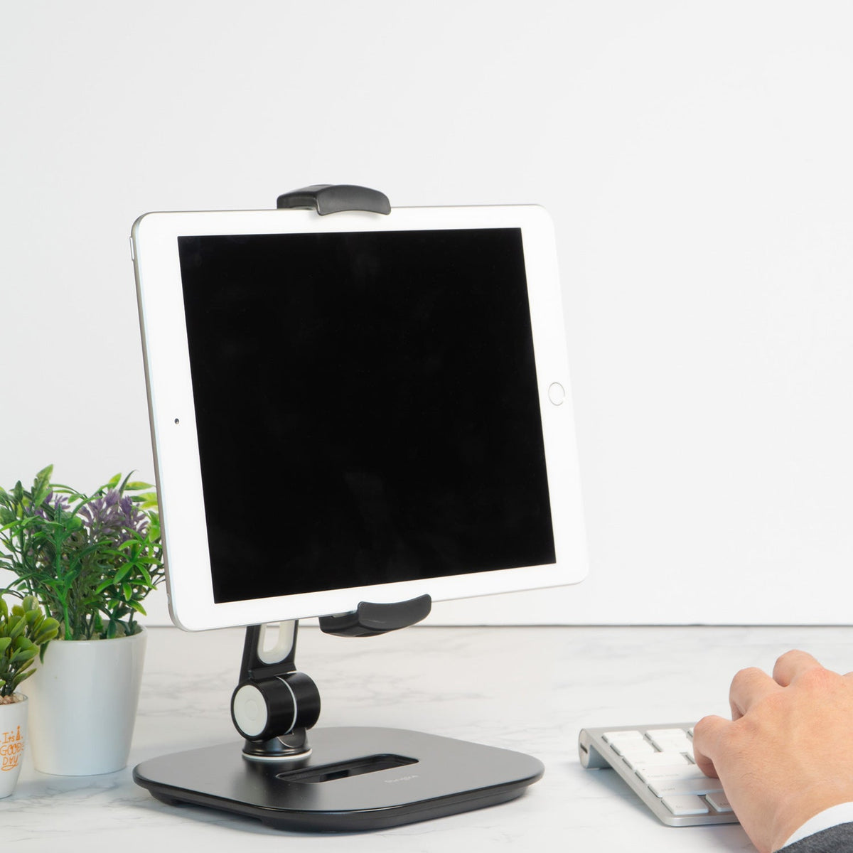 Iron Tablet Stand - A tablet held in a black adjustable stand on a white desk next to a small plant and a computer keyboard.