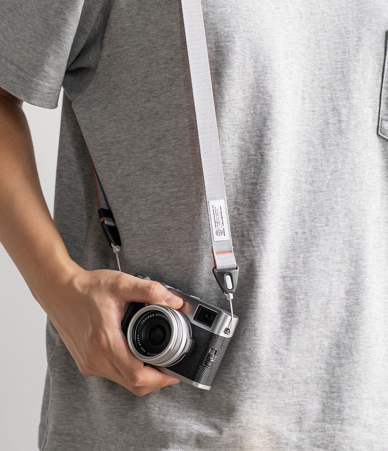 Design Shoulder Strap - Person wearing a light gray camera strap with a small brand tag attached to a vintage-style camera.