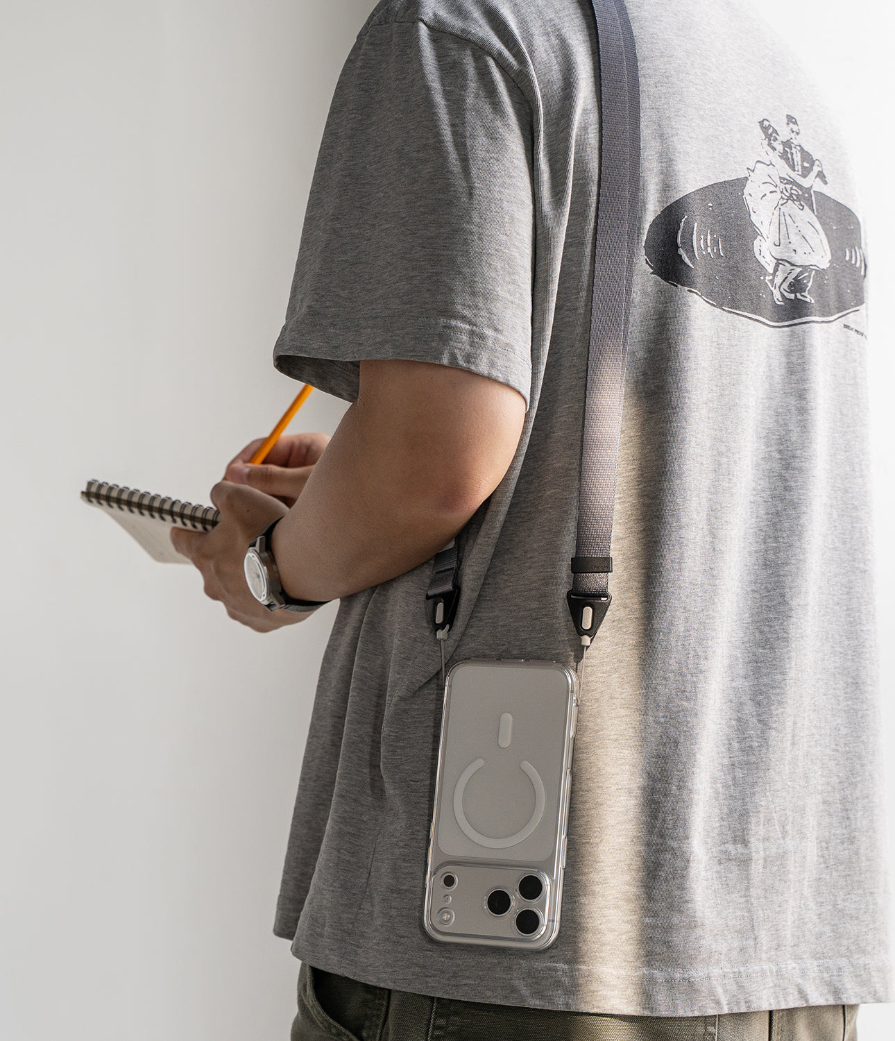 Design Shoulder Strap - A person wearing a grey t-shirt with a phone on a crossbody strap, writing in a notebook.