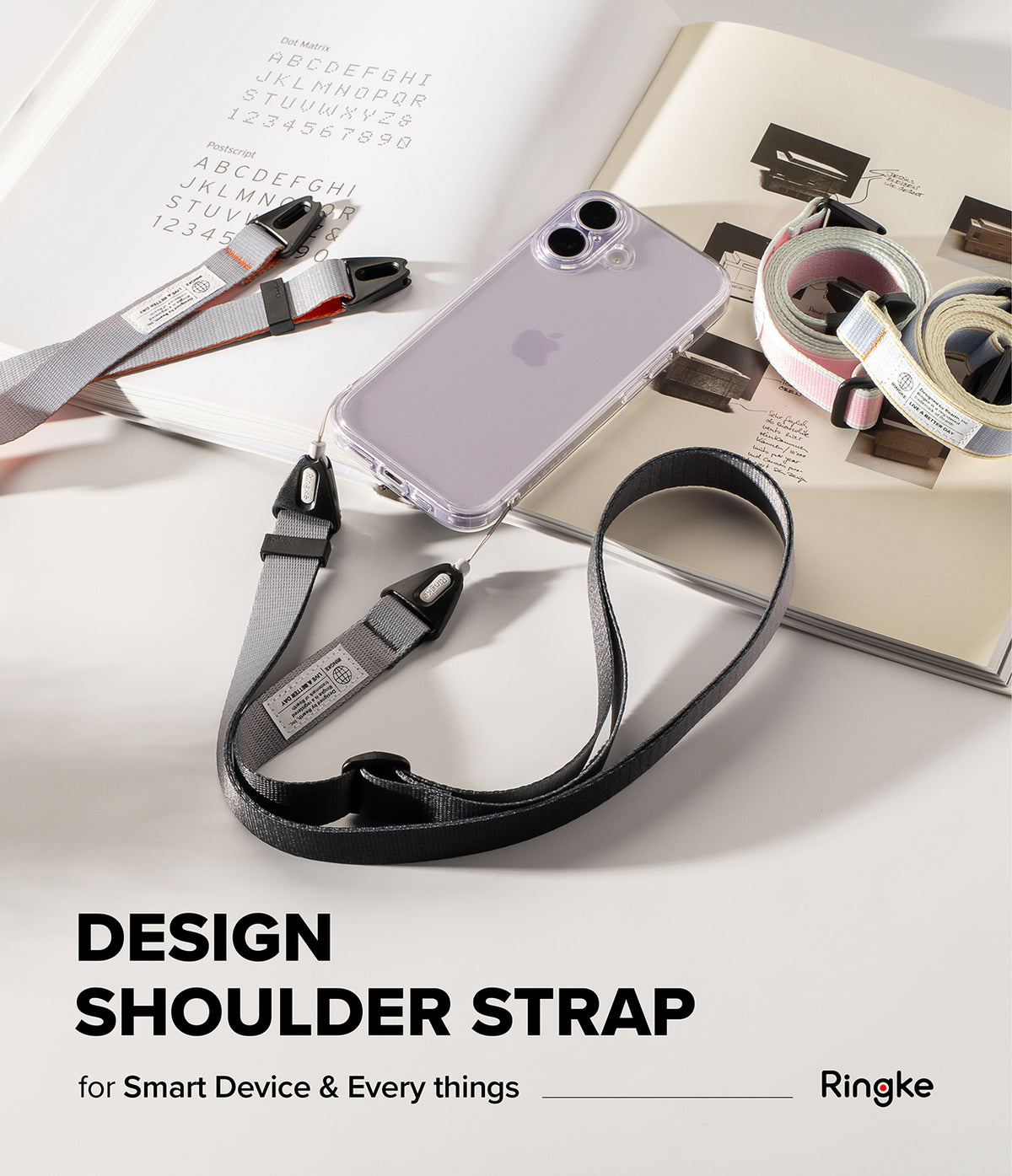 Design Shoulder Strap - A clear phone case with a black shoulder strap resting on an open book with other colorful straps.