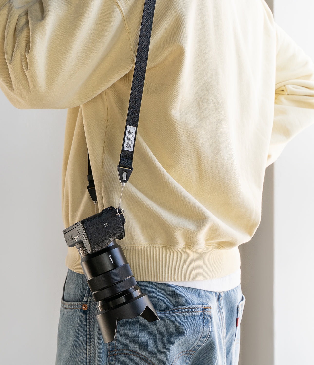 Design Shoulder Strap - Person wearing a black camera strap with a white logo tag attached to a Sony camera over a beige sweater.