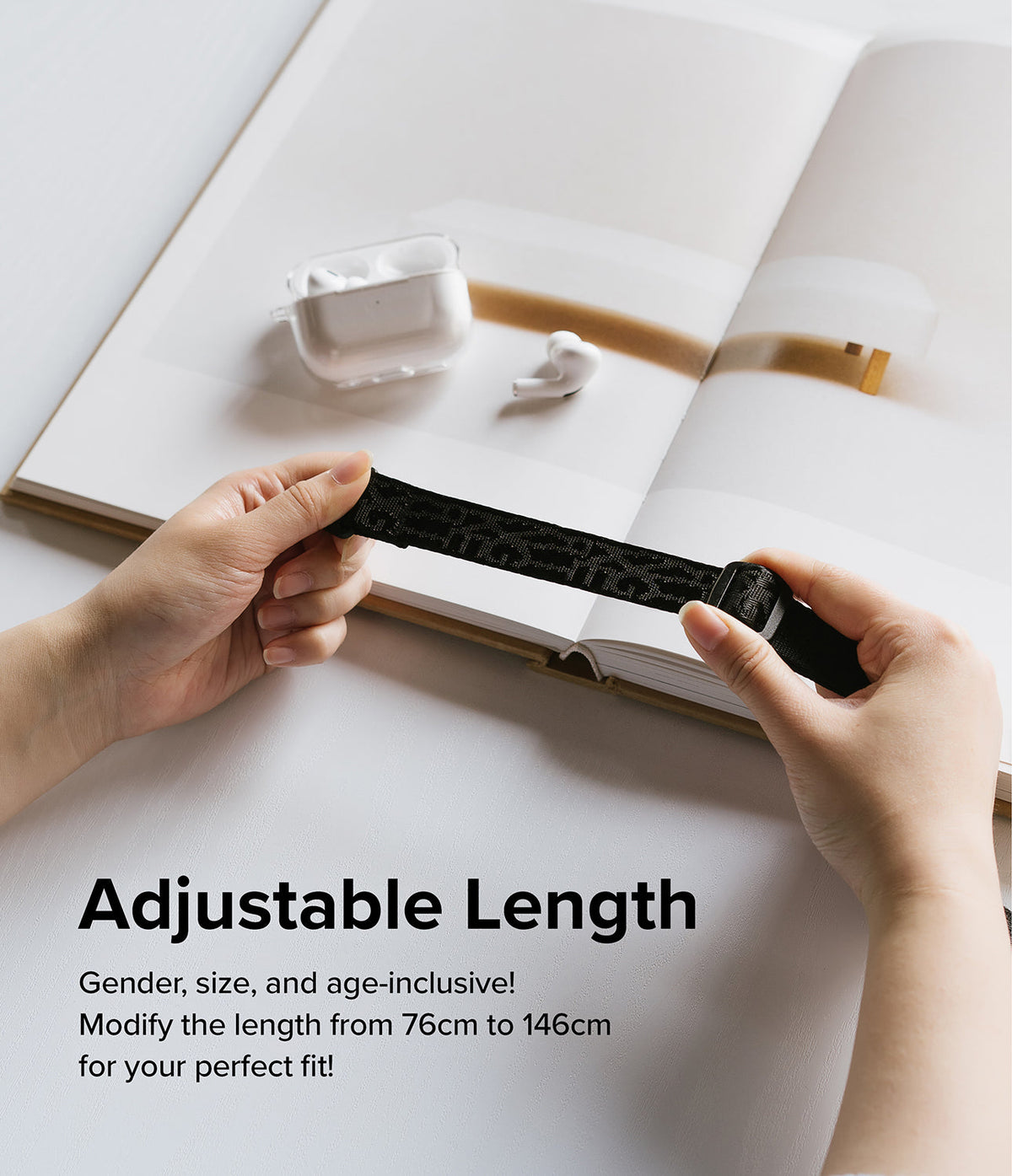 Design Shoulder Strap - Hands adjusting the length of a black patterned strap over an open book with wireless earbuds.