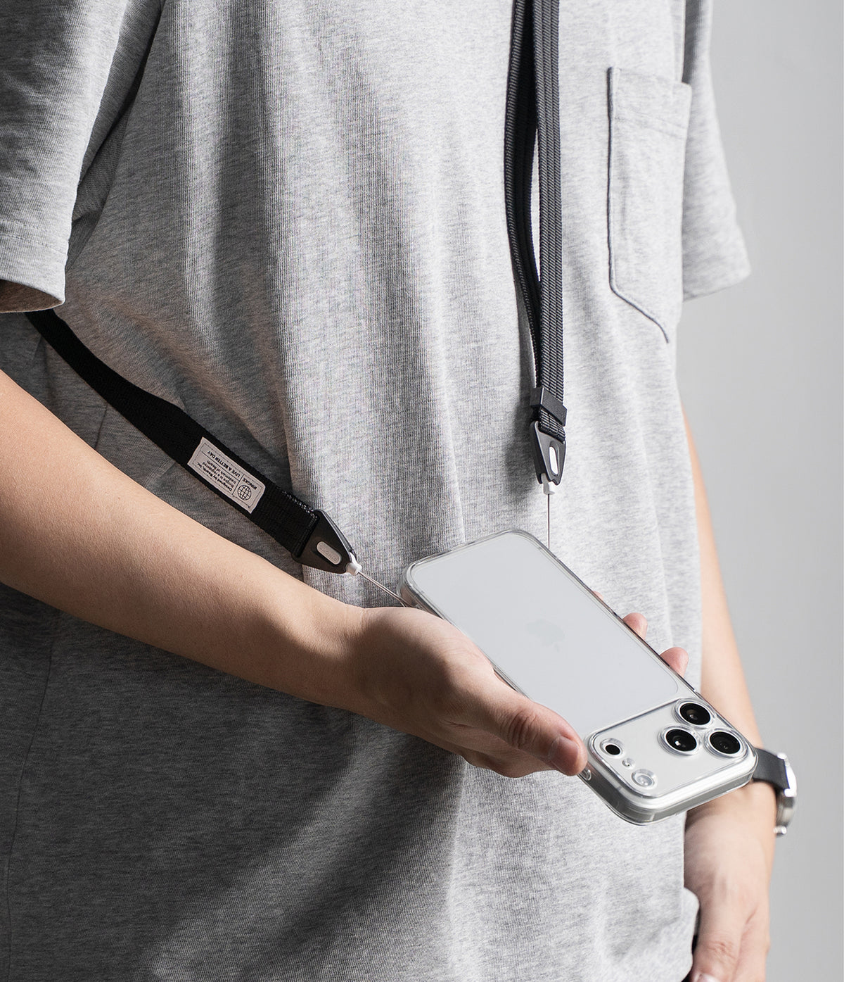 Design Shoulder Strap - A close-up of a person wearing a light gray heather t-shirt and holding a silver smartphone with a black lanyard strap draped around the torso.