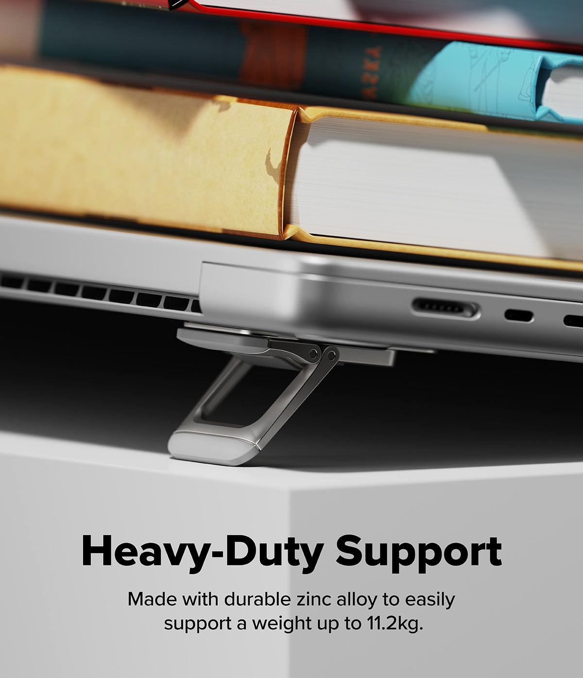 Step-Fit Mini Stand - A close-up of a silver laptop stand supporting a silver laptop, with books stacked on top in a blurred background. Text at the bottom reads 'Heavy-Duty Support' and 'Made with durable zinc alloy to easily support a weight up to 11.