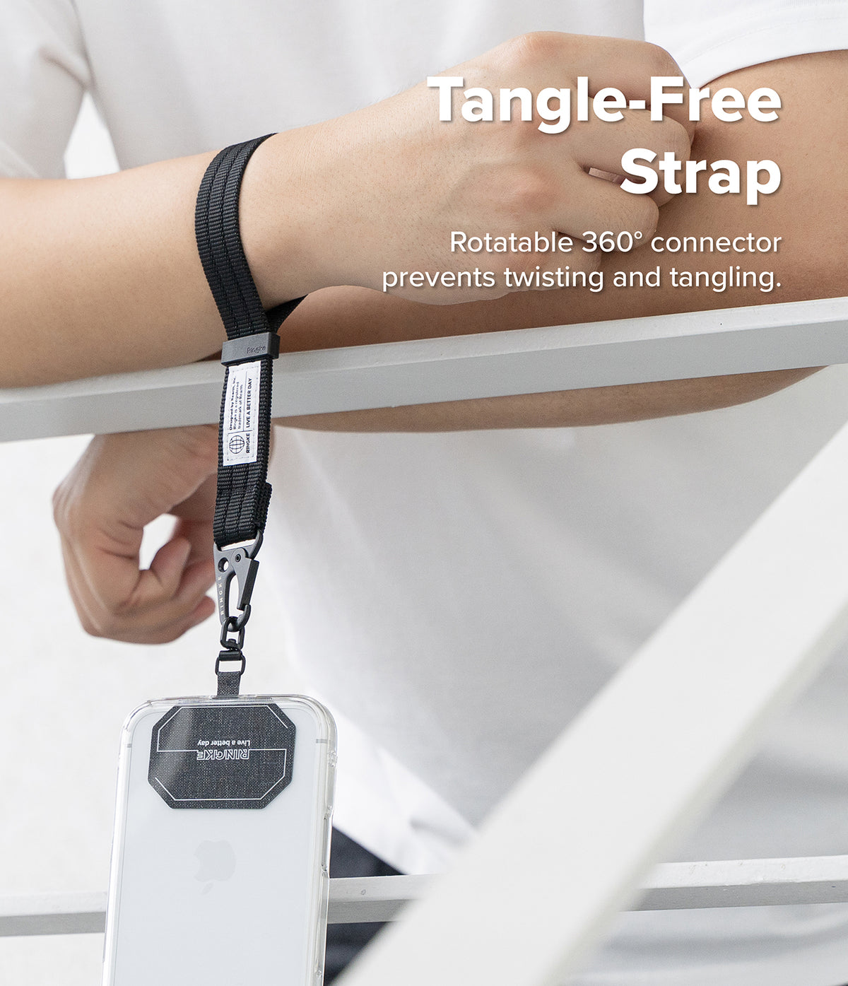 Holder Link Tactical Webbing Hand Strap - A person wearing a black wrist strap lanyard with a phone case attached, shown in a clean indoor setting with white furniture. White text on the image reads 'Tangle-Free Strap' and 'Rotatable 360° connector prevents twisting and tangling.