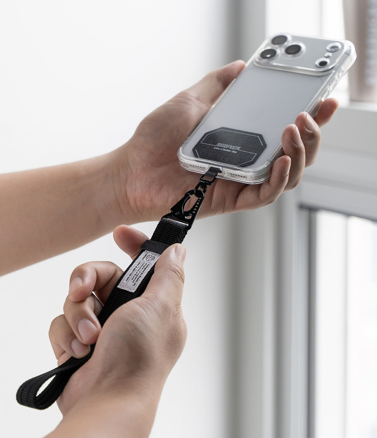 Holder Link Tactical Webbing Hand Strap - A clear phone case with a black magnetic ring and a black wrist strap attached, held by two hands against a bright, softly blurred indoor background.