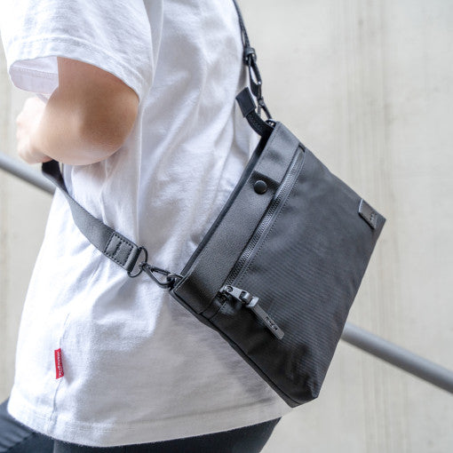 2-Way Bag - Person wearing a black minimalist crossbody bag with a zippered front pocket and adjustable shoulder strap.