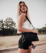 2-Way Bag - Blonde woman wearing a white top and black shorts carries a sleek black crossbody bag outdoors.