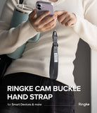 Cam Buckle Hand Strap - Person holding a phone with a Ringke cam buckle hand strap, wearing a beige sweater and black pants.
