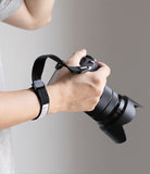 Cam Buckle Hand Strap - A close-up of a person's hand holding a black DSLR camera with a petal-shaped lens hood and a black wrist strap, shown against a neutral gray background.