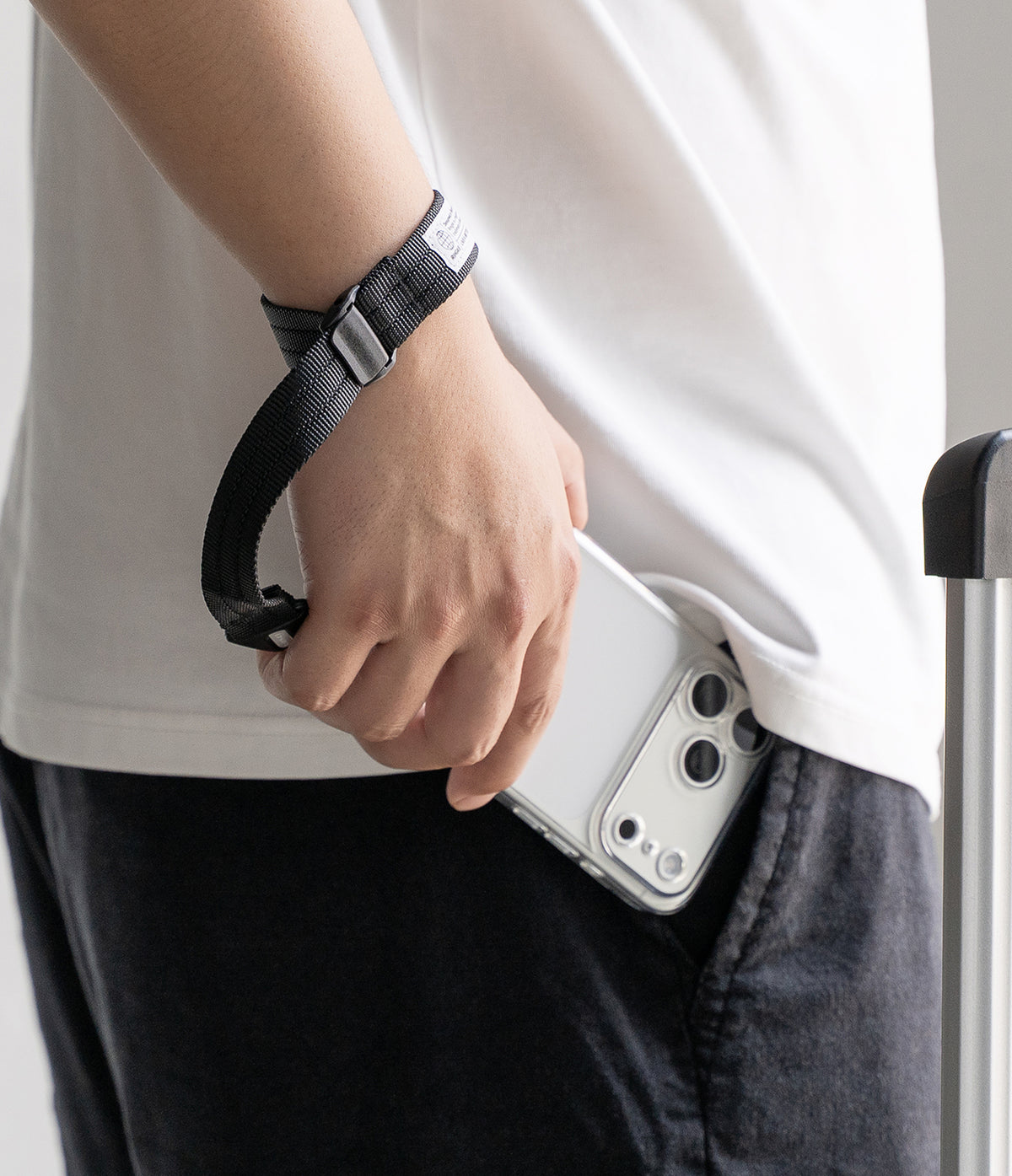 Cam Buckle Hand Strap - A close-up of a person wearing a black wrist strap bag with a silver buckle, holding a white smartphone in their pocket.