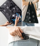 Day-Me Bag - Black foldable tote with pixel art graphics, shown being packed, carried, and stored in a pocket.