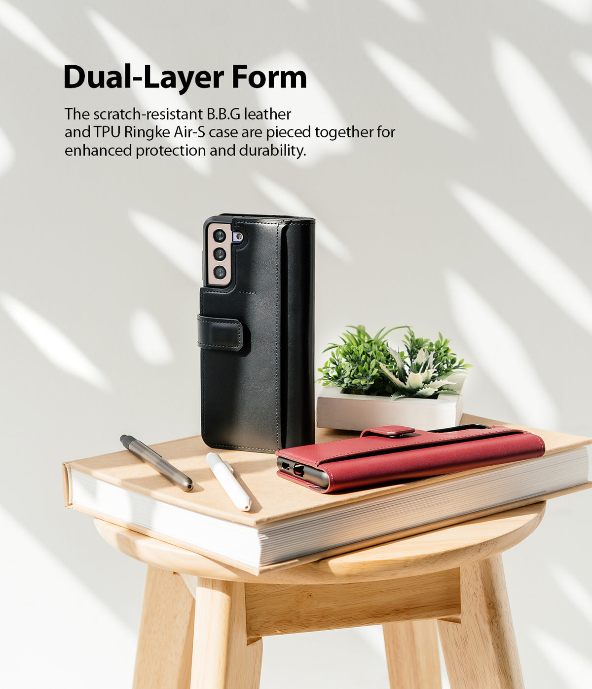 Galaxy S21 Case | Folio Signature+ - Black and red leather folio cases on a book with pens and a small plant on a stool.