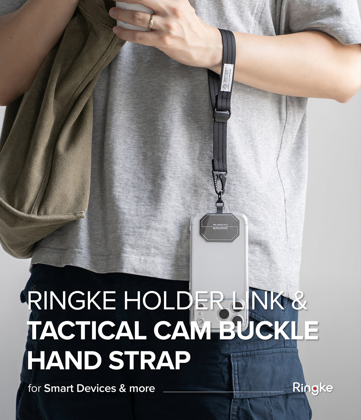 Holder Link Tactical Cam Buckle Hand Strap - A person wearing a gray t-shirt and dark pants holds a beige jacket while wearing a black wrist strap with a clear phone holder and metal buckle. Large white text reads 'RINGKE HOLDER LINK & TACTICAL CAM BUCKLE HAND STRAP' with the Ringke logo at the bottom right.
