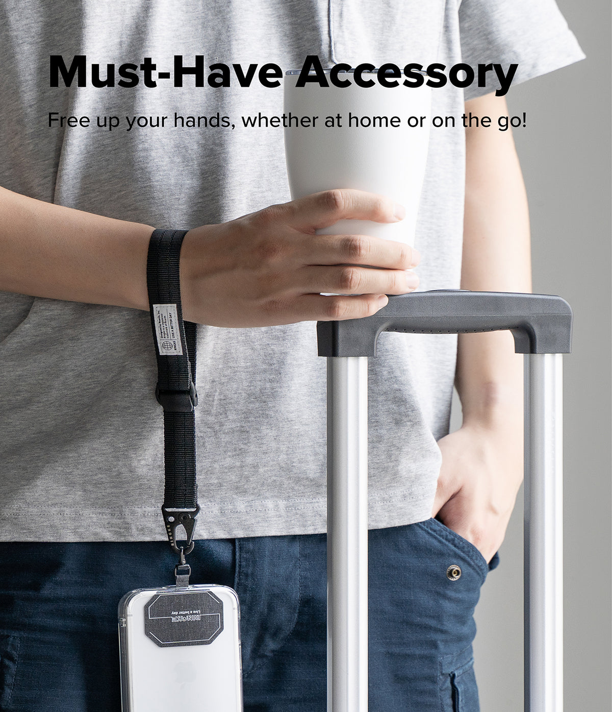 Holder Link Tactical Cam Buckle Hand Strap - A person wearing a gray t-shirt and blue jeans holds a white cup next to a rolling suitcase, with a black wrist strap accessory hanging from the suitcase handle. The text at the top reads 'Must-Have Accessory' and 'Free up your hands, whether at home or on the go!'.