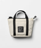 Mini Pouch | Tote Bag - Cream canvas mini tote bag with black handles, zipper, and a square branded patch on the front.