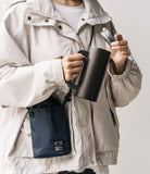Mini Cross Bag | Bucket Bag - Person wearing a beige jacket and a navy blue bucket bag, holding a dark travel mug.