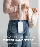 Holder Link Puffer | Hand - A person wearing a beige puffer wrist strap attached to a clear phone case hanging against blue jeans.