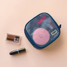 Travel Organizer Pouch - A blue mesh pouch containing a pink compact, next to an eyeshadow palette and a black lipstick.