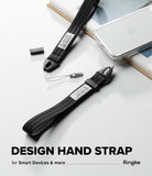 Hand Strap | Basic - A black woven hand strap with a metal clip and a small attached lanyard tag, shown alongside a smartphone and books on a light gray surface. Large text at the bottom reads 'DESIGN HAND STRAP' with smaller text 'for Smart Device & Every things' and the brand name 'Ringke' at the lower right.