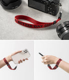 Hand Strap | Lettering - Red fabric wrist strap with black text attached to a camera and a smartphone held in a hand.