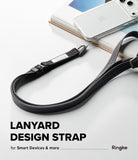 Design Lanyard Strap - A black adjustable lanyard design strap with white stitching and a silver metal clip, shown draped over stacked books with a white smartphone resting on top. Text at the bottom reads 'LANYARD DESIGN STRAP for Smart Devices & more Ringke'.