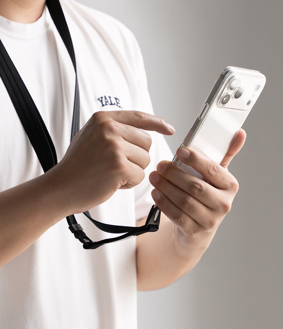 Design Lanyard Strap - A close-up of a person wearing a white t-shirt with a black neck strap and using a white smartphone, shown from the chest up against a neutral gray background.