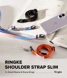 Shoulder Slim Strap - Black, orange, and gradient phone straps arranged on a desk next to a smartphone and an open notebook.