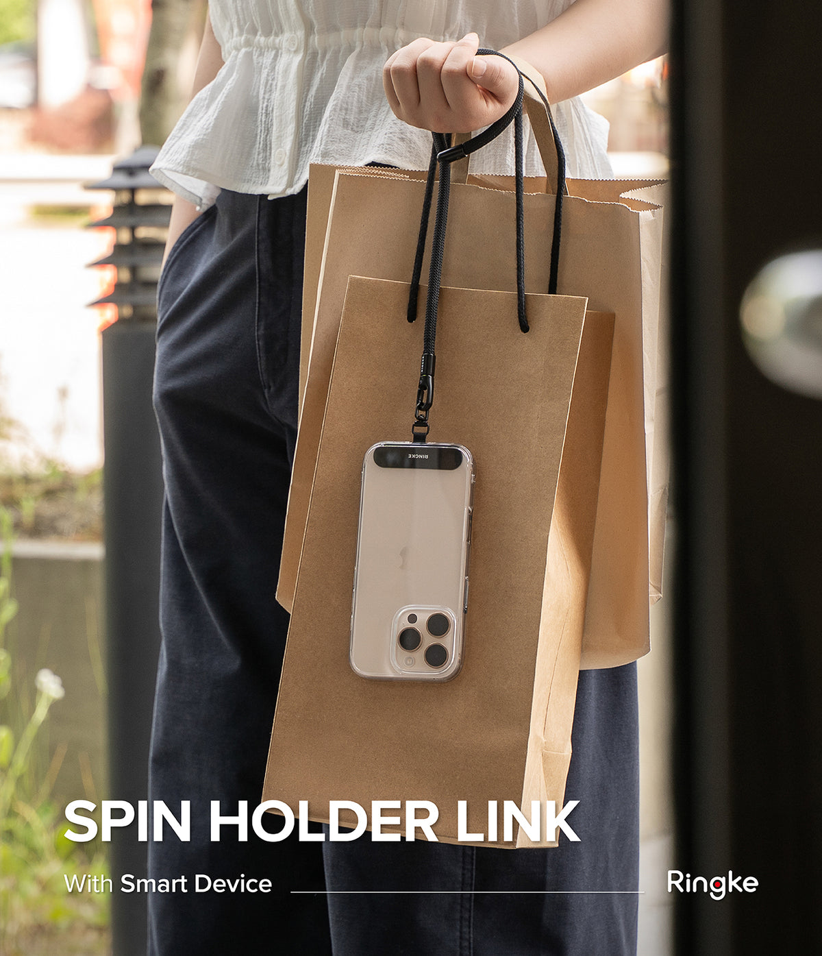 Holder Link Spin - A smartphone in a clear case hangs from a black lanyard held by a person carrying brown paper bags.