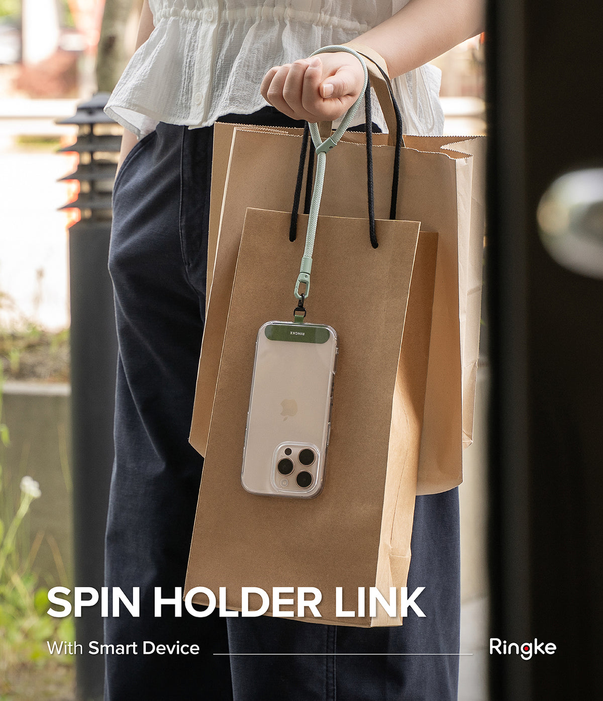 Holder Link Spin - A person holding brown paper shopping bags and a smartphone attached to a sage green wrist lanyard.