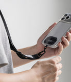 Holder Link Tactical Webbing Lanyard Strap - A close-up of a person holding a smartphone in a clear case with a black wrist strap attached near the camera cutout. The person is wearing a light gray T-shirt, and the background is a clean, neutral light gray studio backdrop.